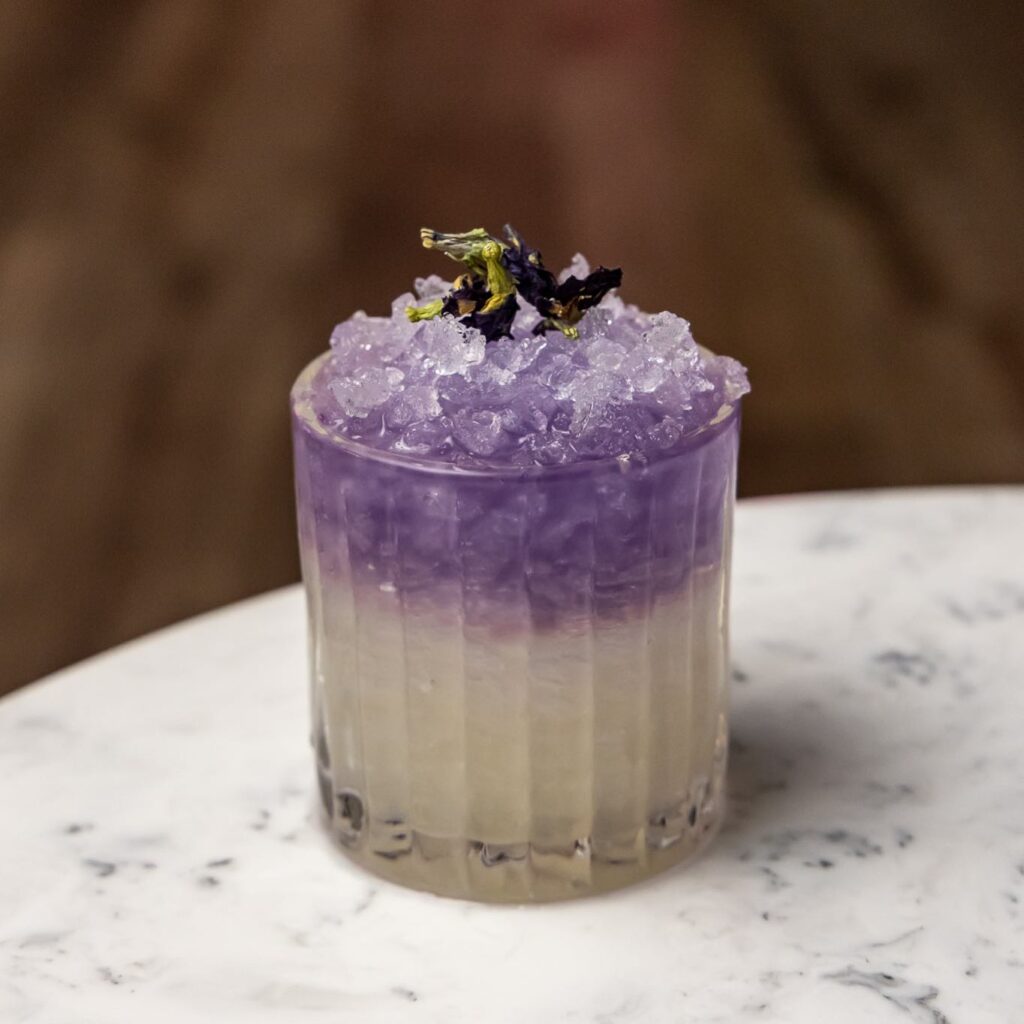 A white and purple cocktail with crushed ice sits on a white marble table